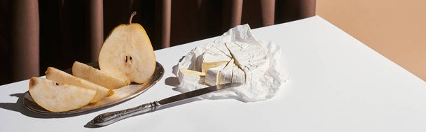 classic still life with pear, brie cheese and knife on table near curtain isolated on beige, panoramic shot - Photo, Image