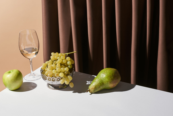 κλασική νεκρή φύση με λευκό κρασί και φρούτα στο τραπέζι κοντά κουρτίνα απομονώνονται σε μπεζ - Φωτογραφία, εικόνα