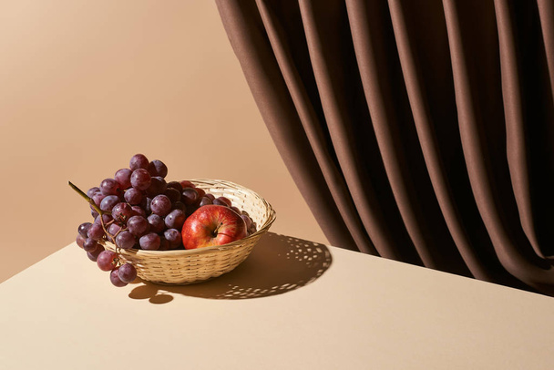 classic still life with pomegranate and grape in wicker basket on table near curtain isolated on beige - Photo, image