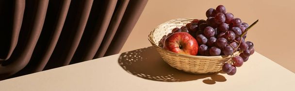 classic still life with pomegranate and grape in wicker basket on table near curtain isolated on beige, panoramic shot - Zdjęcie, obraz