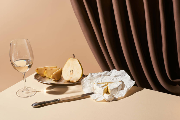 classic still life with pear, white wine and Camembert cheese on table near curtain isolated on beige - Photo, Image
