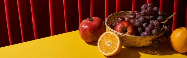 classic still life with fruits in wicker basket on yellow table near red curtain, panoramic shot - Photo, Image