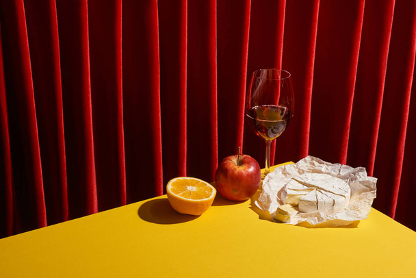 klasická zátiší s Camembert v blízkosti skla červeného vína a ovoce na žlutém stole v blízkosti červené opony - Fotografie, Obrázek