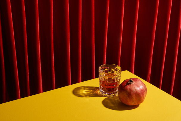Kırmızı perdenin yanındaki sarı masada duran kırmızı şarap kadehinin yanında narlı klasik natürmort. - Fotoğraf, Görsel