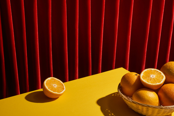 klasická zátiší s pomeranči v proutěném koši na žlutém stole poblíž červené opony - Fotografie, Obrázek