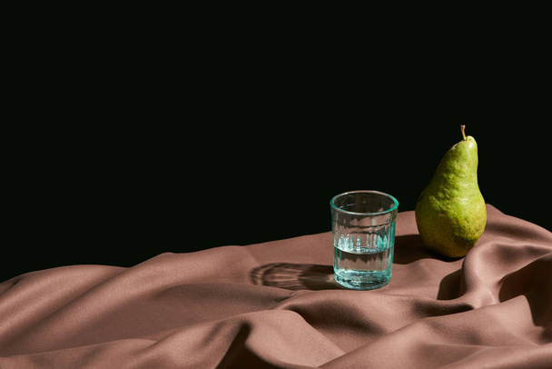 klasyczna martwa natura z gruszką i wodą w szkle na stole z brązowym obrusem wyizolowanym na czarno - Zdjęcie, obraz