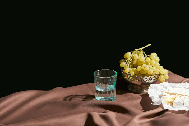 classic still life with Camembert cheese, grape and water in glass on table with brown tablecloth isolated on black - Photo, Image