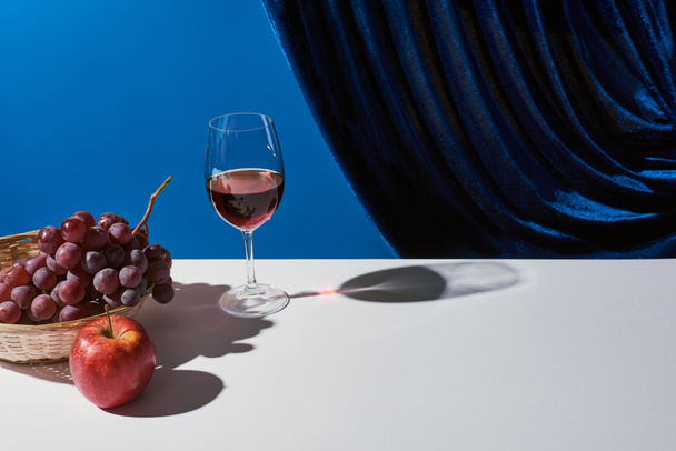 klassiek stilleven met fruit, rode wijn op witte tafel bij velours gordijn geïsoleerd op blauw - Foto, afbeelding