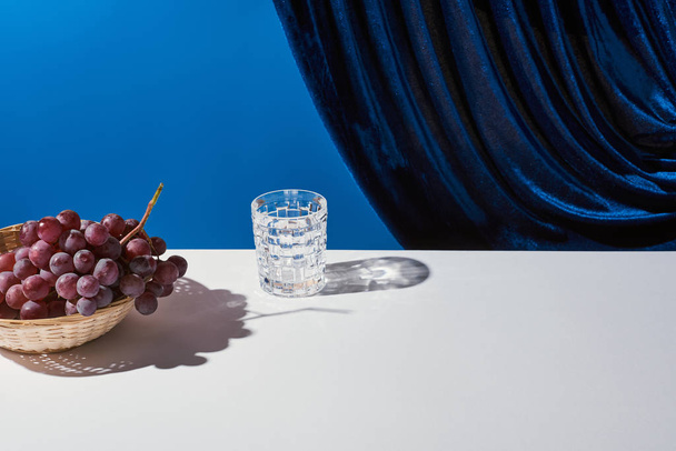 ブルーに隔離されたベロアカーテンの近くの白いテーブルの上のブドウと古典的な静物画 - 写真・画像