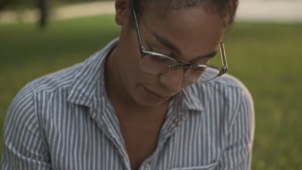 Mosolygó csinos afrikai nő szemüvegben véget vet a beszélgetésnek, és ír valamit utána, miközben ül a parkban - Felvétel, videó