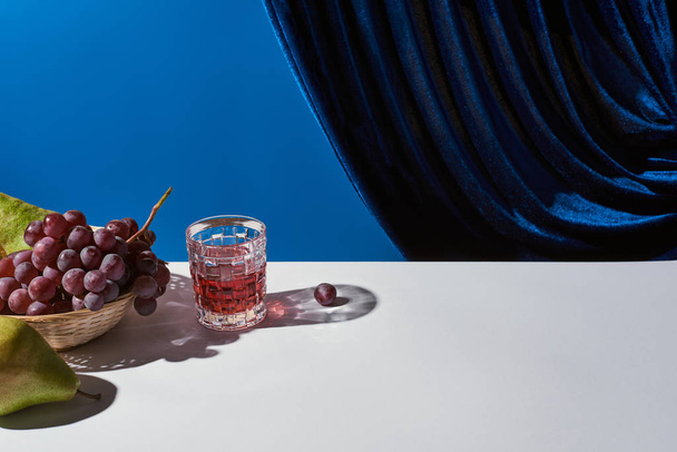 κλασικό νεκρή φύση με σταφύλι, ποτήρι με ποτό σε λευκό τραπέζι κοντά βελούδινη κουρτίνα που απομονώνονται σε μπλε - Φωτογραφία, εικόνα