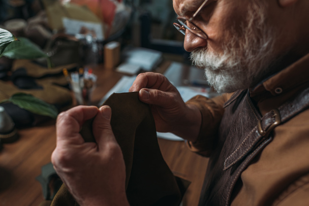 atencioso, sapateiro sênior segurando pedaço de couro genuíno na oficina
 - Foto, Imagem