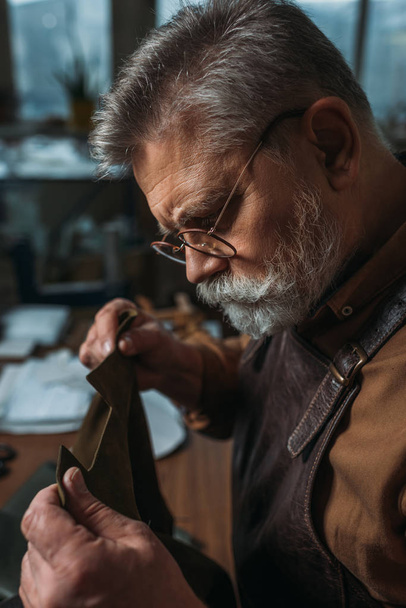 Senior, bärtiger Schuhmacher hält Stück echtes Leder in der Werkstatt - Foto, Bild
