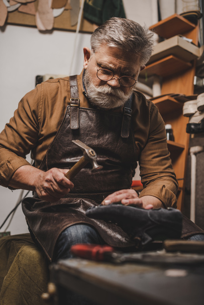 senior, bearded shoemaker holding shoe while repairing shoe in workshop - Photo, Image