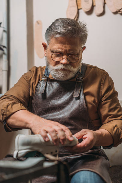 senior, bearded cobbler in leather apron repairing shoe in workshop - Photo, Image