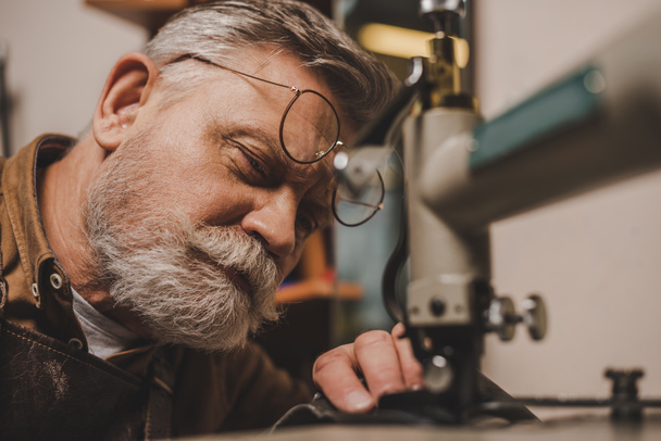 enfoque selectivo de barbudo, zapatero senior de costura de cuero en la máquina de coser en el taller
 - Foto, Imagen