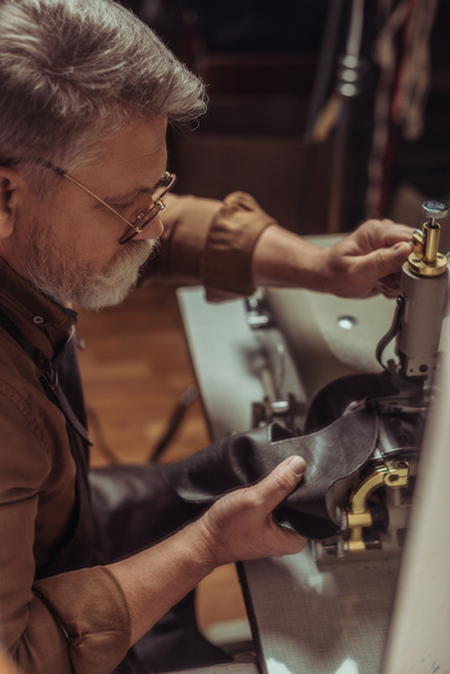 selectieve focus van senior schoenmaker naaien leer op naaimachine in werkplaats - Foto, afbeelding