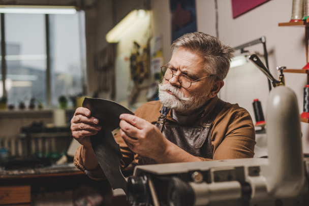 senior, attentive cobbler holding piece of genuine leather near sewing machine - Photo, Image