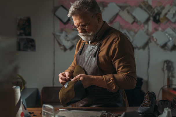 selective focus of senior cobbler holding piece of genuine leather in workshop - Photo, Image