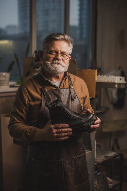 positive, senior shoemaker smiling at camera while holding leather boot - Photo, Image