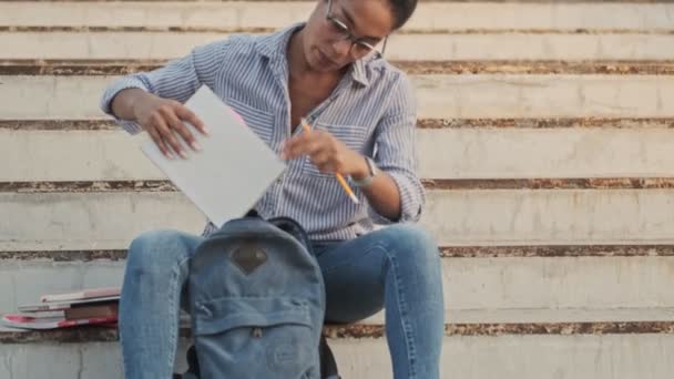 Calm pretty african woman in eyeglasses putting a notebook in a backpack while sitting on stairs outdoors - Felvétel, videó