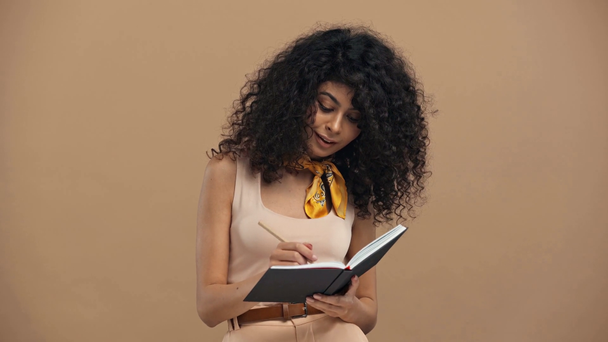 thoughtful bi-racial woman smiling and writing in notebook isolated on beige - Filmagem, Vídeo