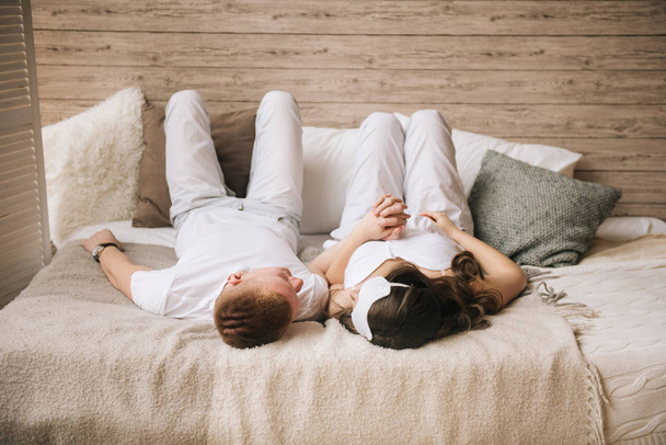 Loving couple in white clothes lying on a light bed holding hands - Foto, afbeelding