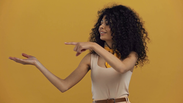sorrindo mulher bi-racial apontando com o dedo para o braço aberto isolado no amarelo escuro
 - Filmagem, Vídeo