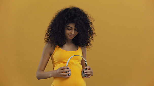 ευτυχισμένη bi-φυλετική έγκυος γυναίκα κρατώντας ακουστικά κοντά στην κοιλιά απομονώνονται σε σκούρο κίτρινο - Πλάνα, βίντεο