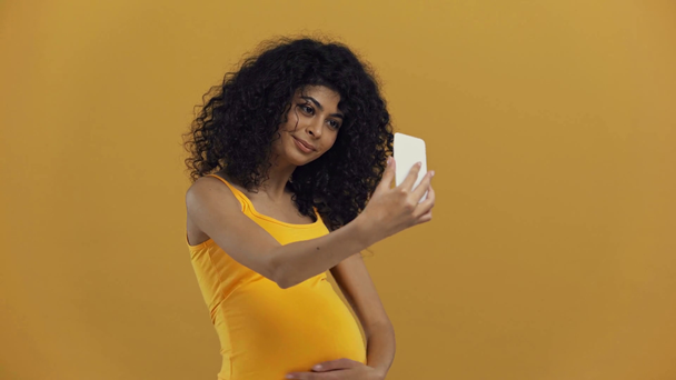 χαμογελαστή αμφι-φυλετική έγκυος γυναίκα που κάνει βιντεοκλήση απομονωμένη στο σκούρο κίτρινο - Πλάνα, βίντεο