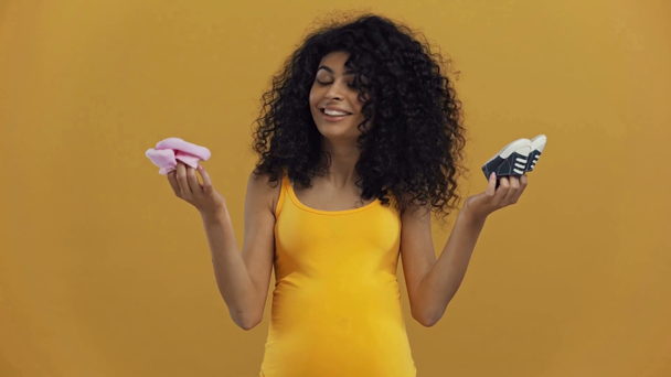 happy bi-racial pregnant woman showing baby booties isolated on dark yellow - Video, Çekim