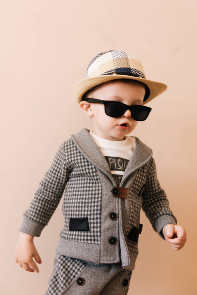 Happy baby boy in grey suit, hat and glasses on light background - 写真・画像