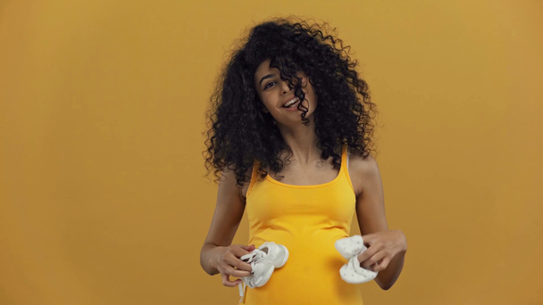 happy bi-racial pregnant woman showing baby booties isolated on dark yellow - Video, Çekim
