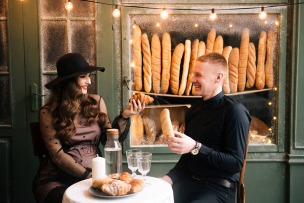 Beautiful young, stylish pregnant girl with her husband sitting at a table with croissants on the background of a French bakery - Фото, зображення