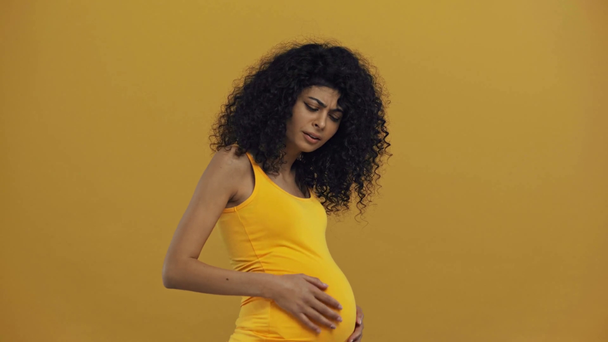 недовольная двурасовая беременная женщина, касающаяся живота, изолированного на темно-желтом
 - Кадры, видео
