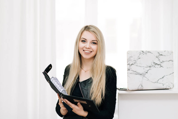 Belle fille blonde souriante avec un cahier et un stylo dans ses mains, à côté d'un ordinateur portable sur un fond clair, employée de bureau
 - Photo, image