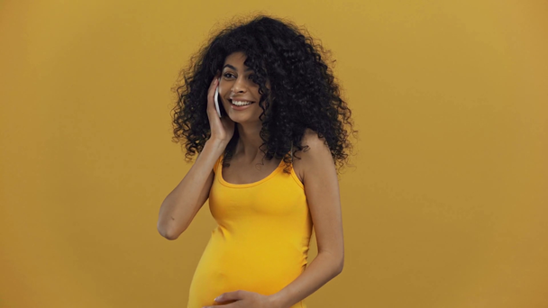 wesoły bi-rasowy kobieta w ciąży rozmawia na smartfonie odizolowany na ciemny żółty - Materiał filmowy, wideo