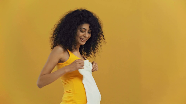 happy bi-racial pregnant woman showing romper isolated on dark yellow - Metraje, vídeo