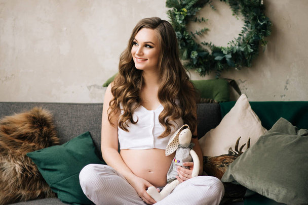 Beautiful young, stylish pregnant girl in white pajamas sitting on the couch in a stylish interior - Valokuva, kuva
