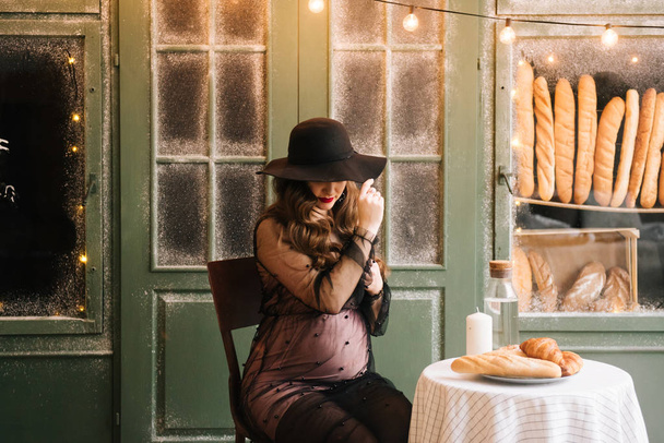 Beautiful young, stylish pregnant girl, in a black dress and hat, in the interior of a French bakery - Foto, Imagem