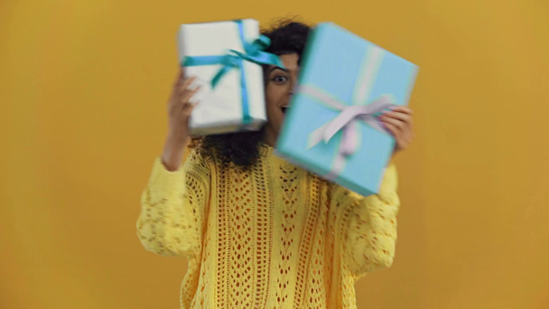 happy bi-racial woman showing gift boxes isolated on dark yellow - Filmmaterial, Video
