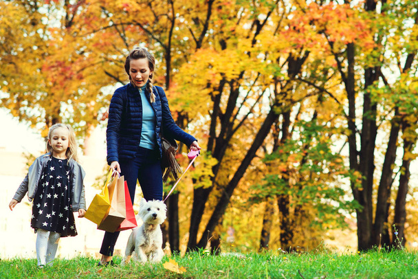 Anne ve kızı alışveriş yaptıktan ve birlikte yürüdükten sonra iyi vakit geçiriyorlar. Sonbahar parkında köpekle yürüyüşe çıkmış bir aile. - Fotoğraf, Görsel