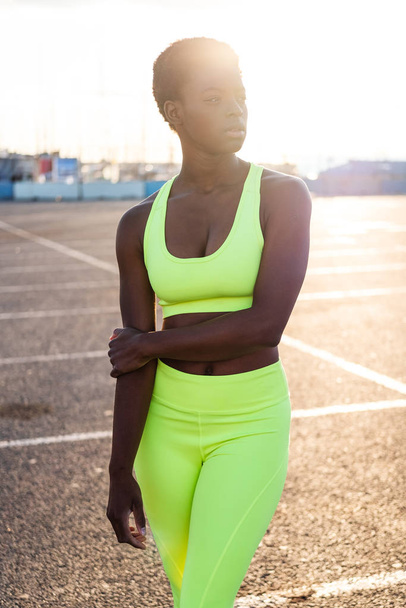 Σοβαρή εστιασμένη Αφρο-Αμερικανίδα γυναίκα με ζωηρά κίτρινα αθλητικά να κοιτάζει αλλού και να σκέφτεται σε ηλιαχτίδες μόνη της στο δρόμο ενάντια στο αστικό περιβάλλον - Φωτογραφία, εικόνα