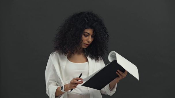 serious bi-racial businesswoman writing on papers isolated on black - Кадри, відео
