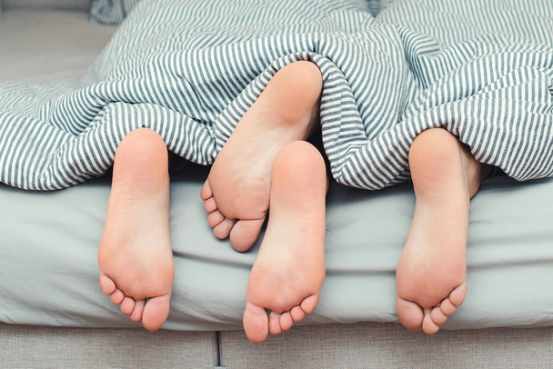 Loving couple is lying on bed under blanket. Cropped image of young couple is lying on bed. Close up of male and female feet - Фото, изображение