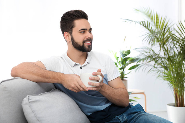Young man with cup of drink relaxing on couch at home - Fotoğraf, Görsel