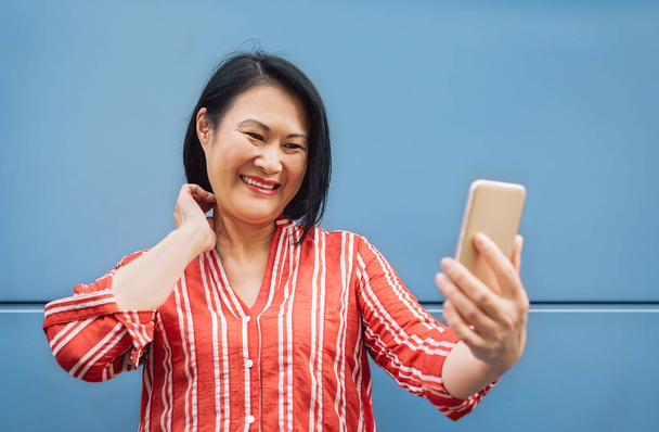 Mulher asiática feliz usando smartphone móvel ao ar livre - Sénior se divertindo com a nova tecnologia da moda para aplicativos de mídia social - Tecnologia de estilo de vida idoso e conceito de pessoas idosas
 - Foto, Imagem