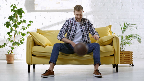 Χαρούμενος άντρας κάθεται στον καναπέ και παίζει με το μπάσκετ - Πλάνα, βίντεο