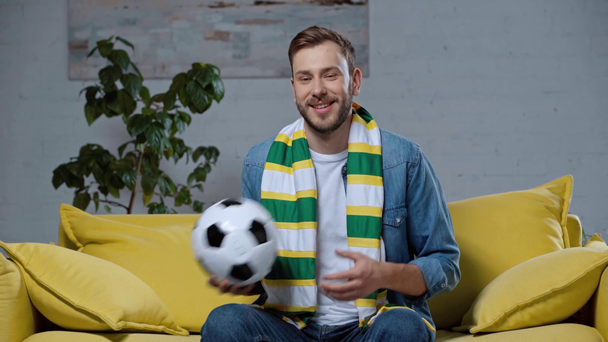 cheerful man sitting on sofa and playing with soccer ball - Кадри, відео