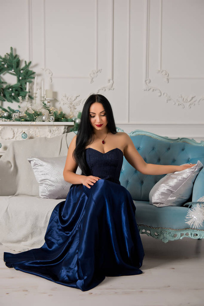Girl with black hair in a blue dress on a background of Christmas decorations. - Фото, изображение